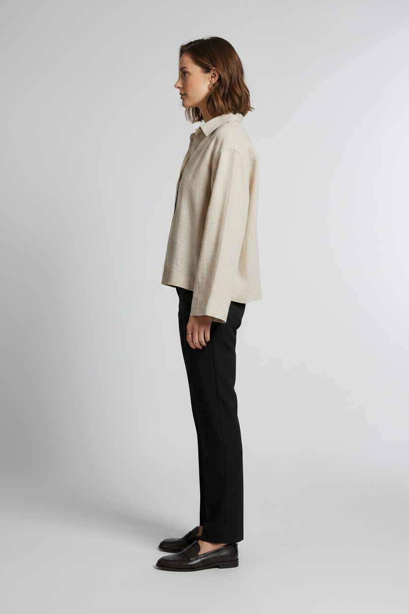 A woman stands against a light gray studio background wearing a light beige linen button-up shirt with long sleeves, black trousers, and black loafers.