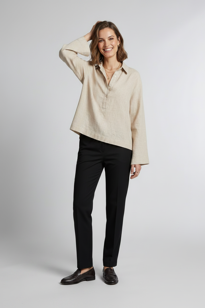 A woman stands against a light gray studio background wearing a light beige linen button-up shirt with long sleeves, black trousers, and black loafers.