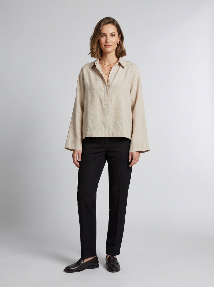 A woman stands against a light gray studio background wearing a light beige linen button-up shirt with long sleeves, black trousers, and black loafers.