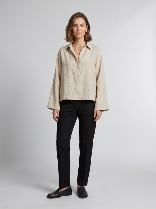 A woman stands against a light gray studio background wearing a light beige linen button-up shirt with long sleeves, black trousers, and black loafers.