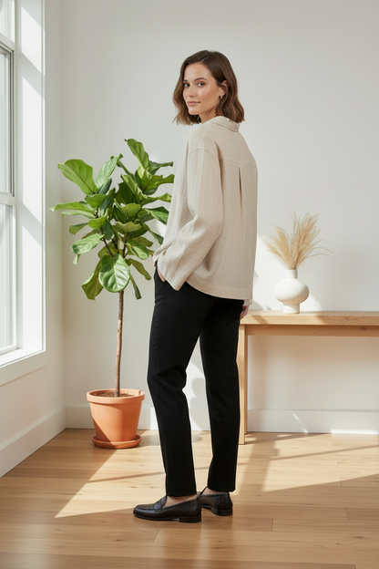 A woman stands in a bright room with wood floors, wearing a light beige linen button-up shirt, black trousers, and black loafers. A potted fiddle-leaf fig and a wooden console table with a vase are in the background.