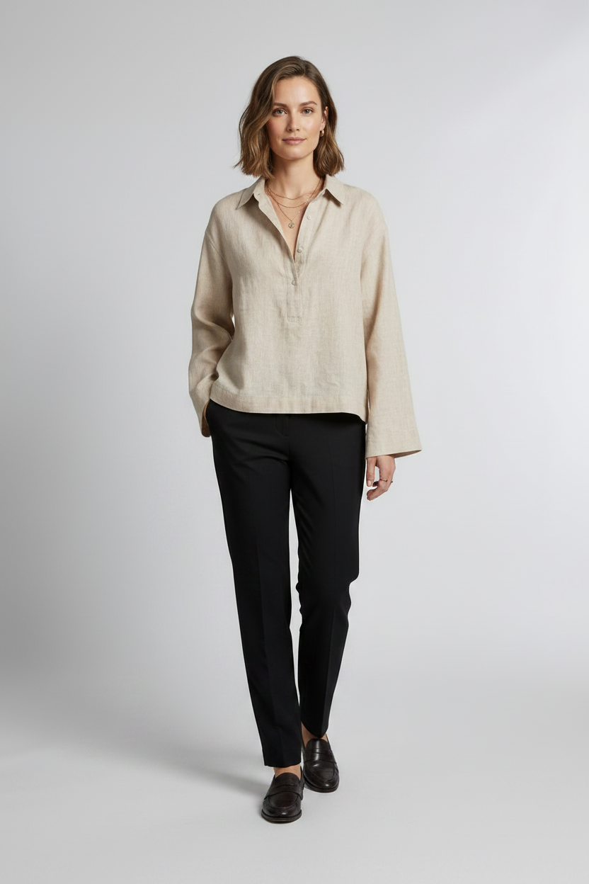 A woman stands against a light gray studio background wearing a light beige linen button-up shirt with long sleeves, black trousers, and black loafers.