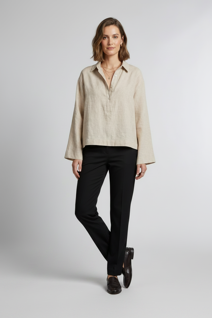 A woman stands against a light gray studio background wearing a light beige linen button-up shirt with long sleeves, black trousers, and black loafers.