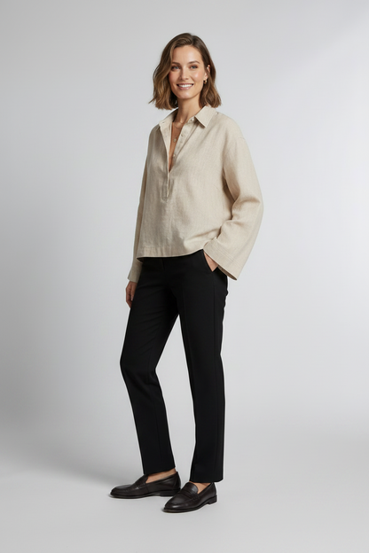 A woman stands against a light gray studio background wearing a light beige linen button-up shirt with long sleeves, black trousers, and black loafers.