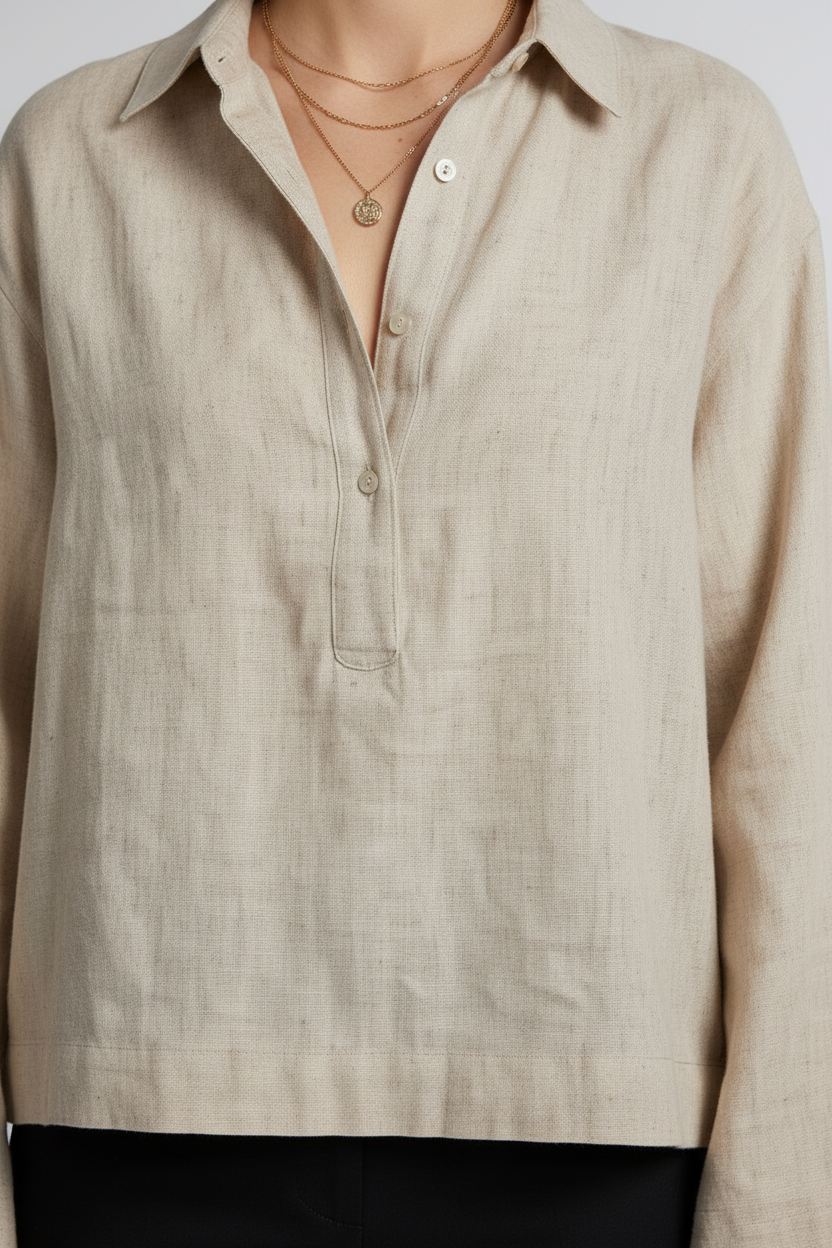 A woman stands against a light gray studio background wearing a light beige linen button-up shirt with long sleeves, black trousers, and black loafers.