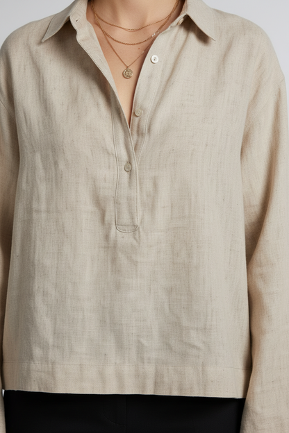 A woman stands against a light gray studio background wearing a light beige linen button-up shirt with long sleeves, black trousers, and black loafers.