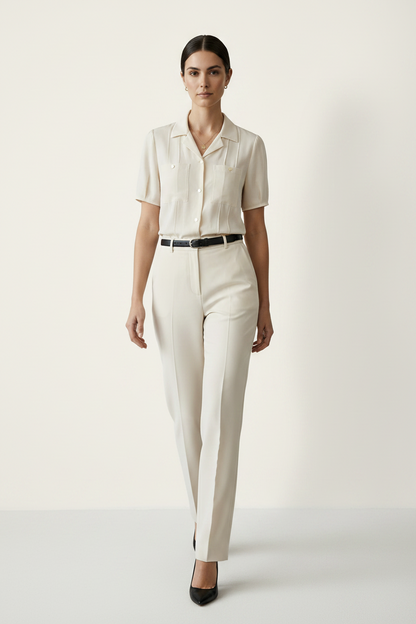 A woman modeling a short-sleeve cream-colored button-up shirt with structured shoulders and chest pockets, paired with matching cream trousers and a black belt, against a plain light beige studio background.