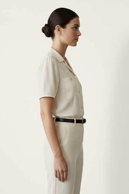 A woman modeling a short-sleeve cream-colored button-up shirt with structured shoulders and chest pockets, paired with matching cream trousers and a black belt, against a plain light beige studio background.