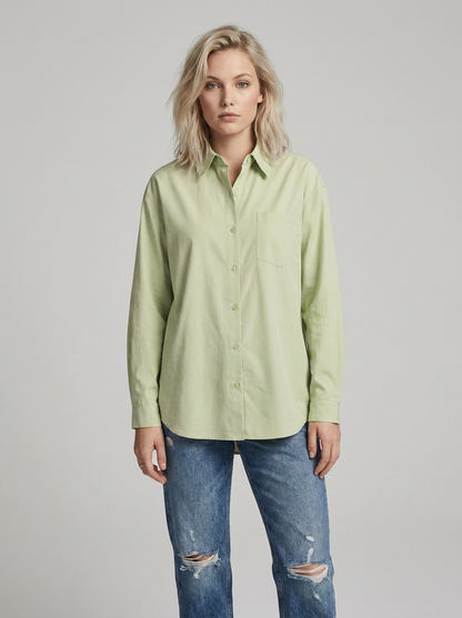 A woman modeling a light sage green long-sleeve button-up shirt with a chest pocket, paired with distressed blue jeans, against a solid light gray studio background.