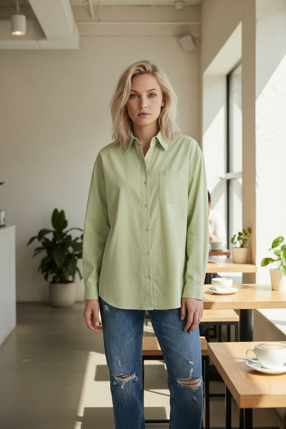 A woman modeling a light sage green long-sleeve button-up shirt with a chest pocket, paired with distressed blue jeans, against a solid light gray studio background.