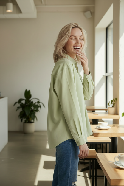 A woman laughing in a bright cafe setting, wearing a light sage green button-up shirt and blue jeans.