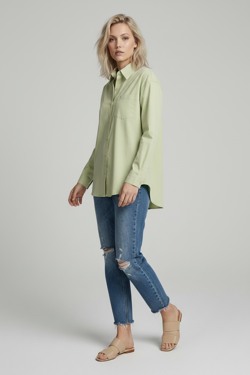 A woman modeling a light sage green long-sleeve button-up shirt with a chest pocket, paired with distressed blue jeans, against a solid light gray studio background.