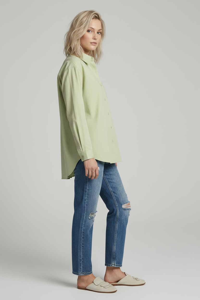 A woman modeling a light sage green long-sleeve button-up shirt with a chest pocket, paired with distressed blue jeans, against a solid light gray studio background.