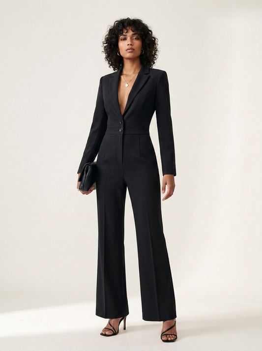 A model wearing a black tailored jumpsuit with a deep V-neckline, single front button, structured shoulders, and wide-leg trousers, styled with black strappy heels and a small black clutch against a plain light background.