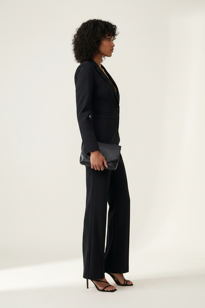 A model wearing a black tailored jumpsuit with a deep V-neckline, single front button, structured shoulders, and wide-leg trousers, styled with black strappy heels and a small black clutch against a plain light background.