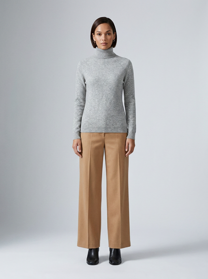 A model stands against a light gray studio background wearing a light gray ribbed turtleneck sweater, camel wide-leg trousers, and black ankle boots.