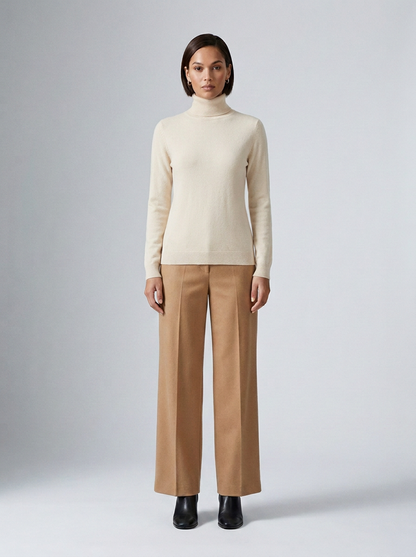 A model stands against a light gray studio background wearing a cream ribbed turtleneck sweater, camel wide-leg trousers, and black ankle boots.