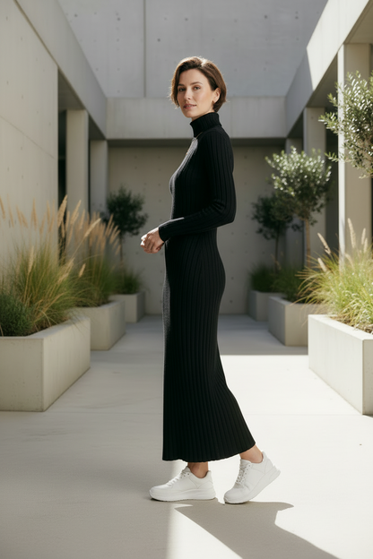 A woman wearing a black ribbed long-sleeve turtleneck maxi dress and white sneakers walks down a modern concrete walkway lined with potted plants.