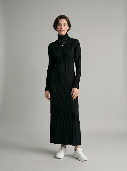 A woman modeling a long-sleeved, black ribbed turtleneck maxi dress with white sneakers, standing against a neutral light gray studio background.