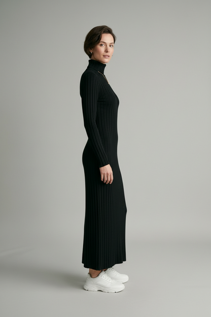 A woman modeling a long-sleeved, black ribbed turtleneck maxi dress with white sneakers, standing against a neutral light gray studio background.