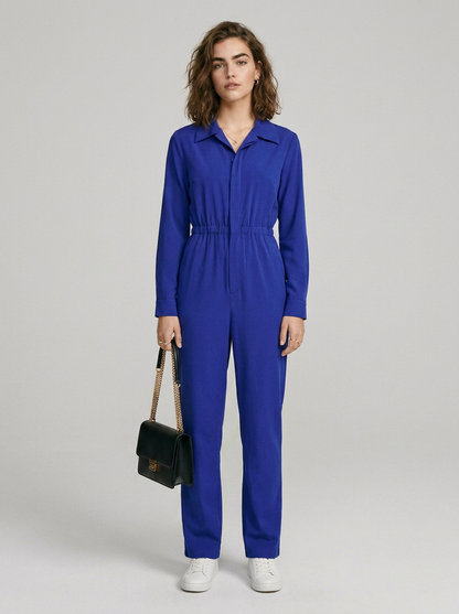 A model stands against a light gray studio background wearing a bright royal blue one-piece jumpsuit with a collar, front zipper, drawstring waist, and straight legs. She is holding a small black shoulder bag and wearing white sneakers.