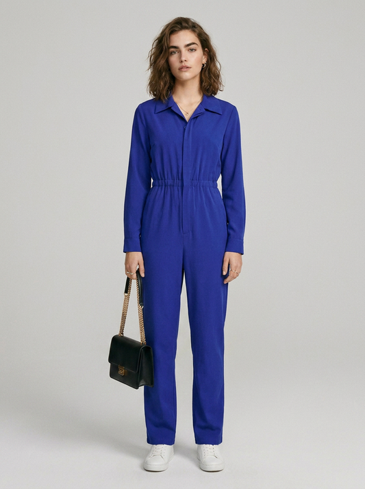 A model stands against a light gray studio background wearing a bright royal blue one-piece jumpsuit with a collar, front zipper, drawstring waist, and straight legs. She is holding a small black shoulder bag and wearing white sneakers.