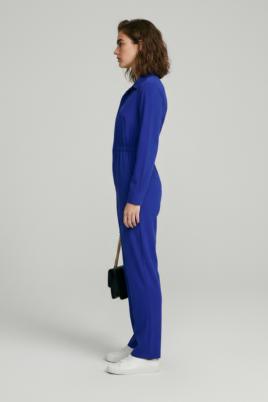 A model stands against a light gray studio background wearing a bright royal blue one-piece jumpsuit with a collar, front zipper, drawstring waist, and straight legs. She is holding a small black shoulder bag and wearing white sneakers.