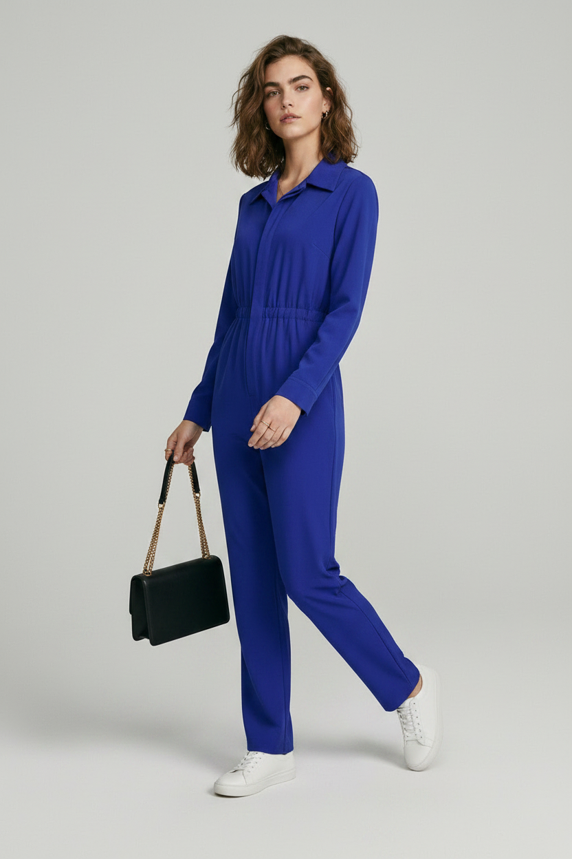 A model stands against a light gray studio background wearing a bright royal blue one-piece jumpsuit with a collar, front zipper, drawstring waist, and straight legs. She is holding a small black shoulder bag and wearing white sneakers.