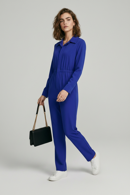 A model stands against a light gray studio background wearing a bright royal blue one-piece jumpsuit with a collar, front zipper, drawstring waist, and straight legs. She is holding a small black shoulder bag and wearing white sneakers.
