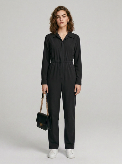A woman stands against a light gray studio background wearing a black long-sleeve jumpsuit with a collar, front zipper, and drawstring waist. She is holding a small black handbag with a gold chain strap and wearing white sneakers.