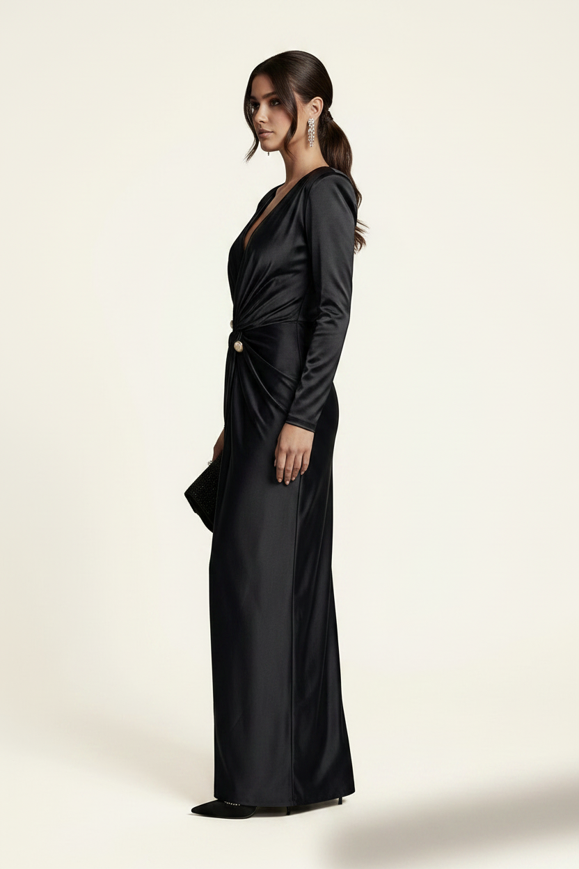 A model wears a floor-length black satin gown with structured shoulders, a deep V-neckline, and a gathered waist detail accented by gold buttons. She is holding a small black clutch against a clean, off-white studio background.