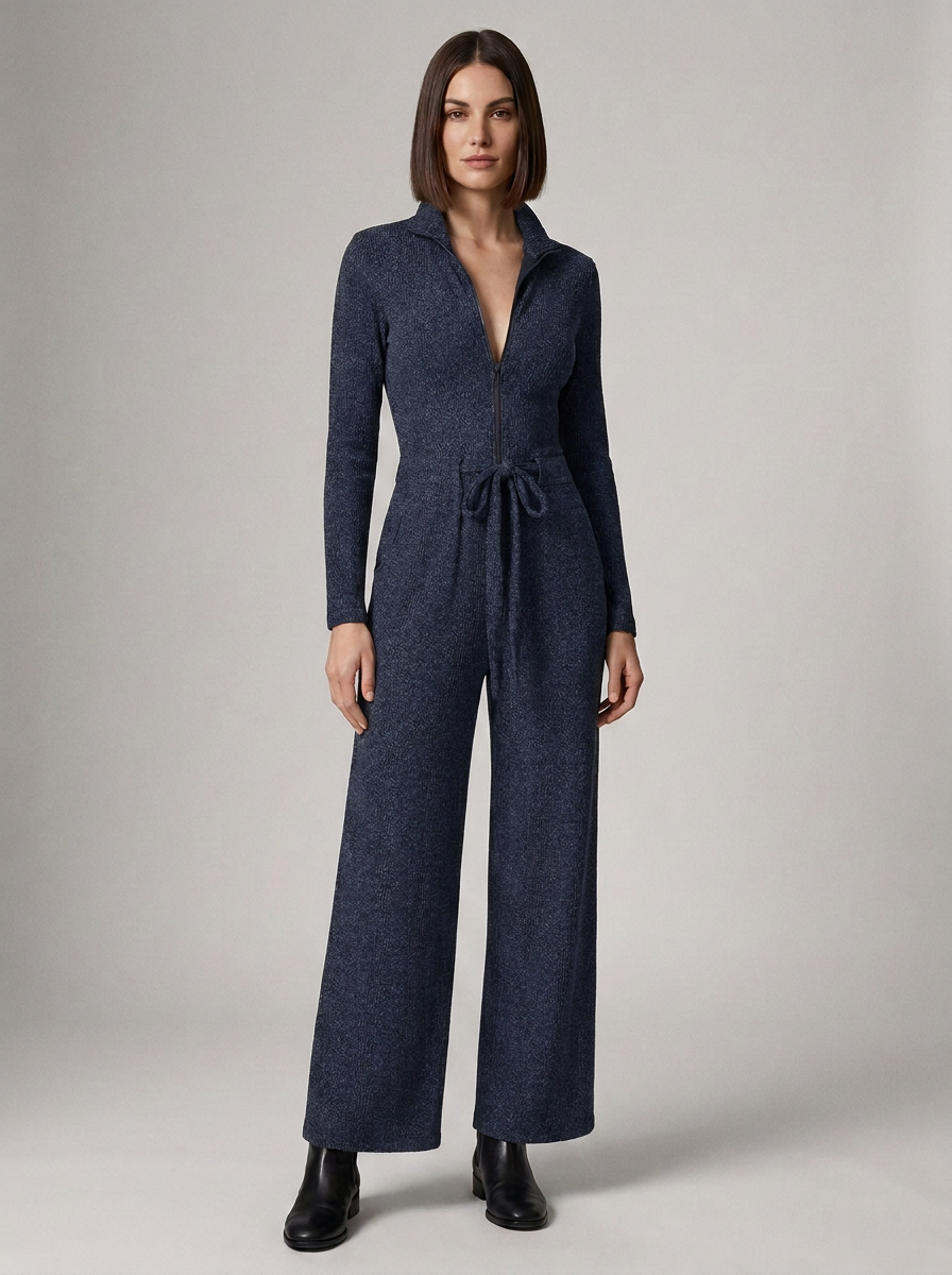 A woman modeling a dark navy blue knit jumpsuit with a collared neckline, front zipper, long sleeves, a tie waist, and wide legs, paired with black ankle boots against a neutral studio background.