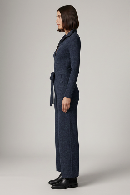 A woman modeling a dark navy blue knit jumpsuit with a collared neckline, front zipper, long sleeves, a tie waist, and wide legs, paired with black ankle boots against a neutral studio background.