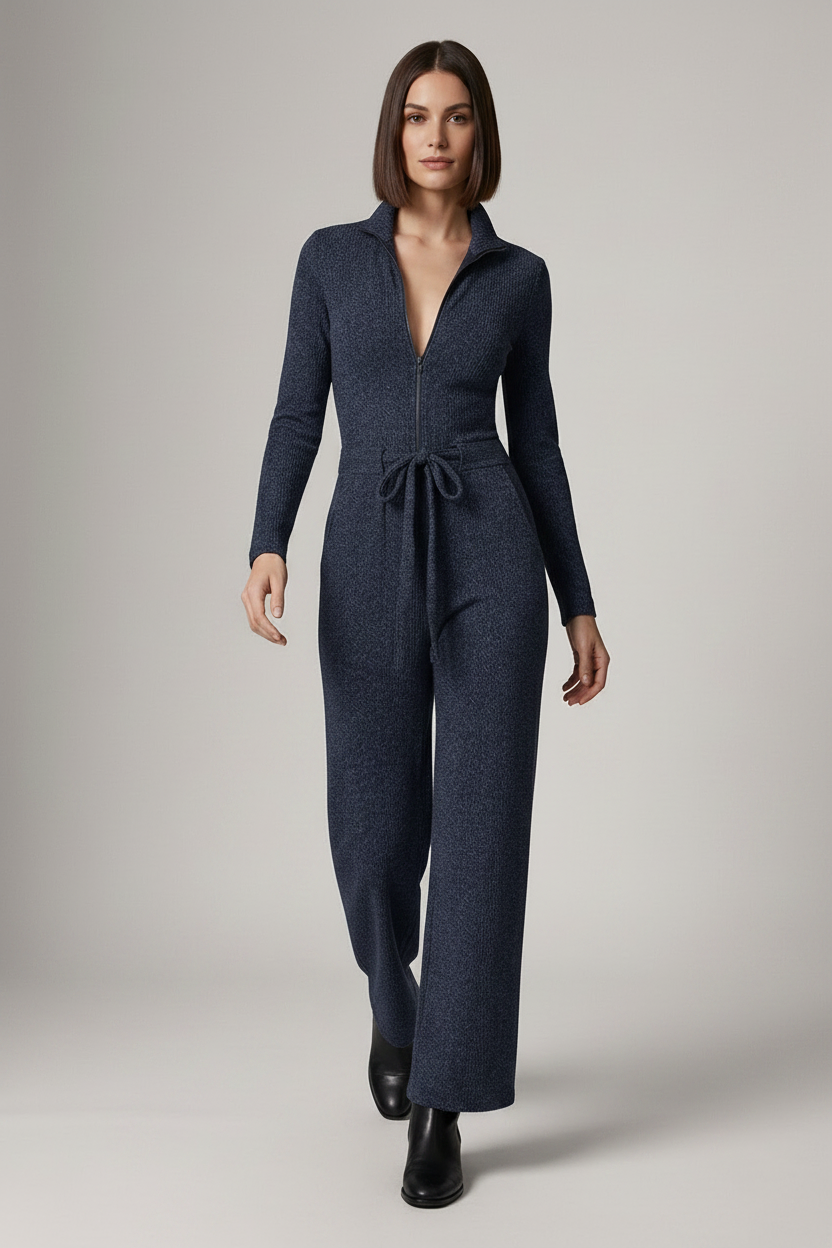 A woman modeling a dark navy blue knit jumpsuit with a collared neckline, front zipper, long sleeves, a tie waist, and wide legs, paired with black ankle boots against a neutral studio background.