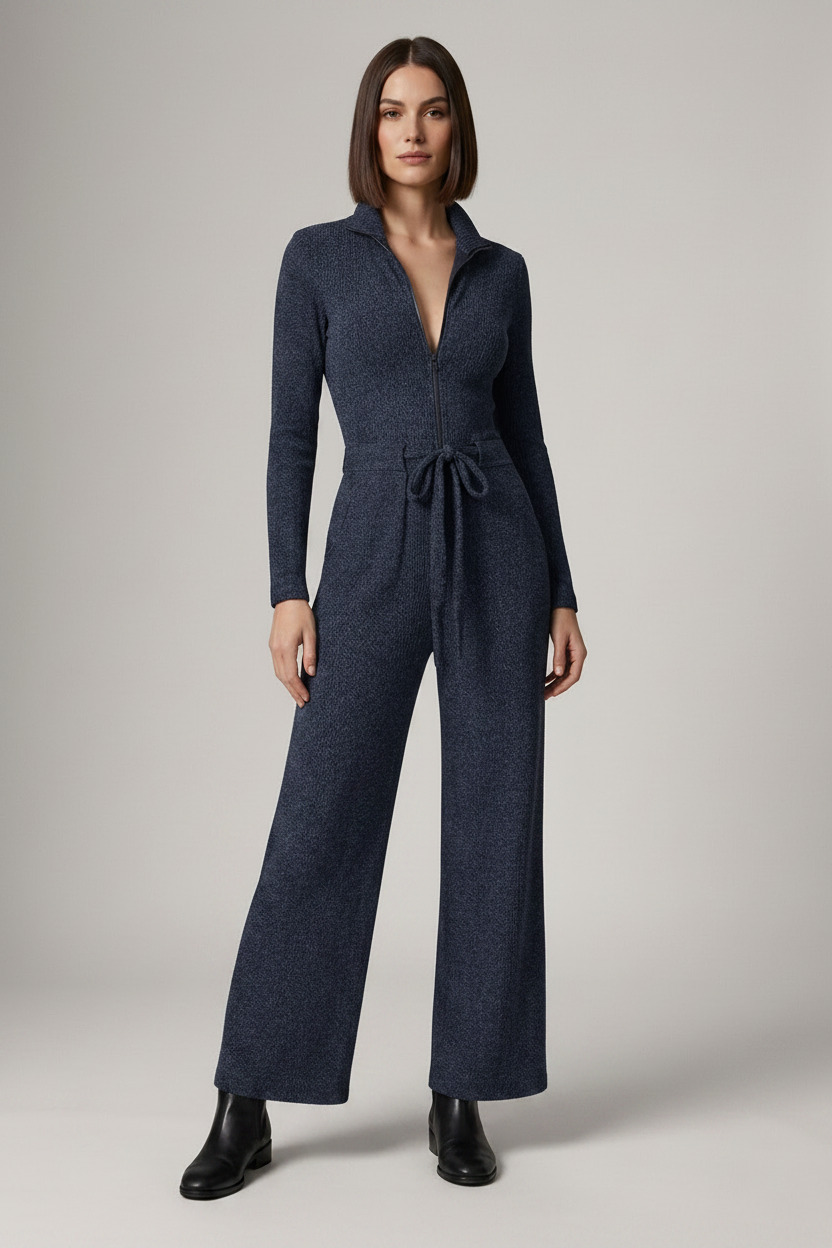 A woman modeling a dark navy blue knit jumpsuit with a collared neckline, front zipper, long sleeves, a tie waist, and wide legs, paired with black ankle boots against a neutral studio background.
