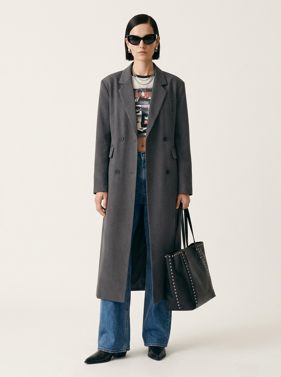 A model stands against a light beige studio background wearing a long charcoal gray double-breasted coat over a graphic tee, blue jeans, black shoes, and holding a black studded tote bag.