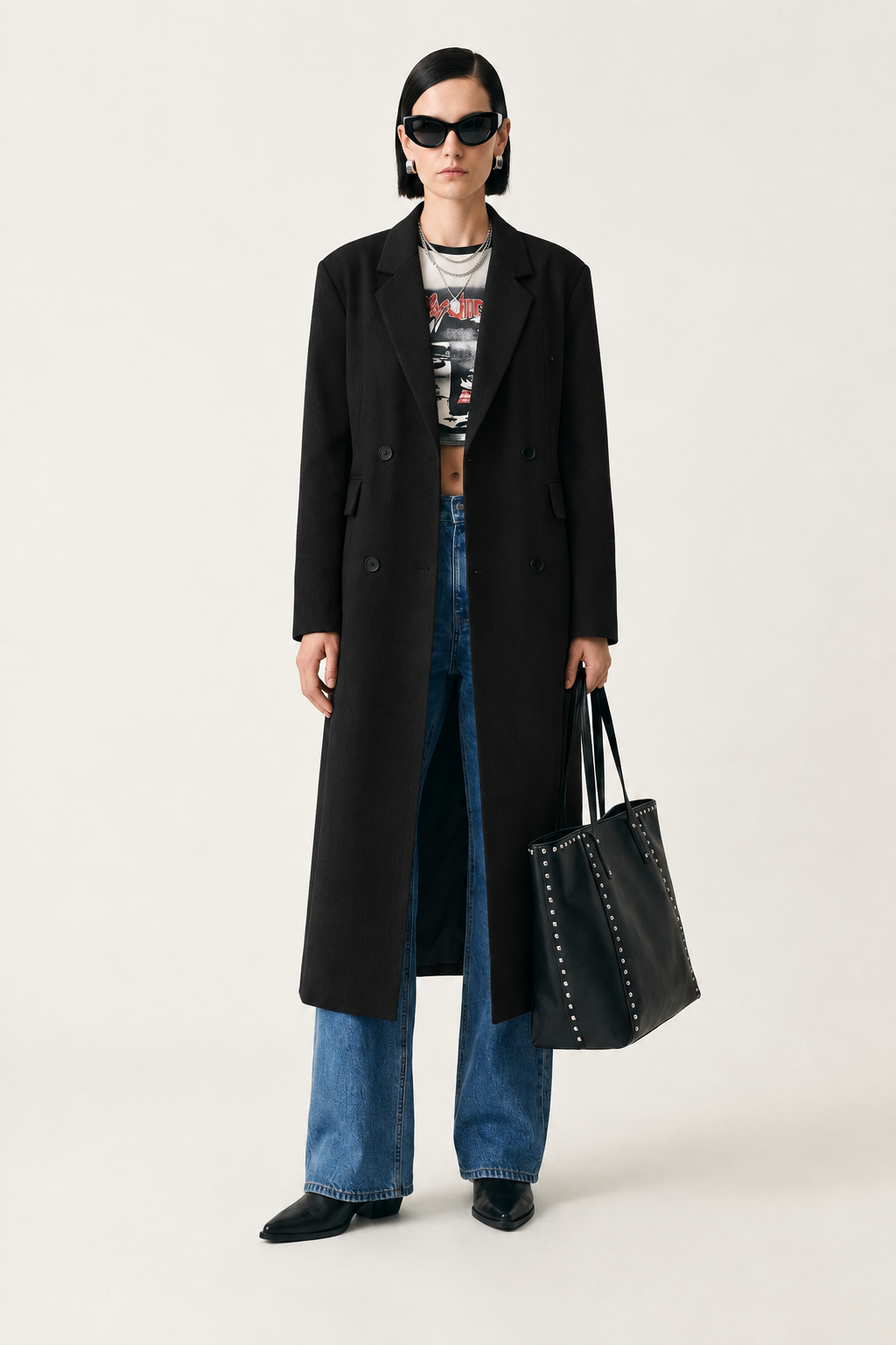 A model stands against a light gray studio background wearing a long black double-breasted coat over a graphic crop top, blue jeans, black pointed shoes, and black sunglasses, while holding a black leather tote bag with silver studs.