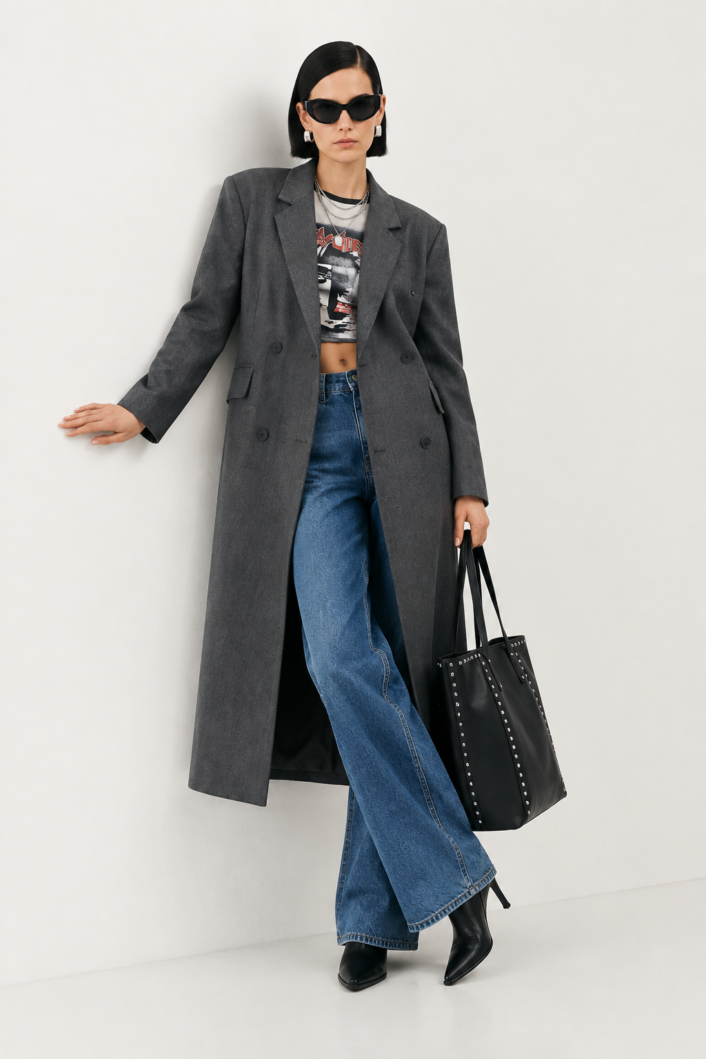A model wearing a long charcoal gray double-breasted overcoat, blue wide-leg jeans, a graphic crop top, and black ankle boots, holding a black studded tote bag against a plain off-white studio background.