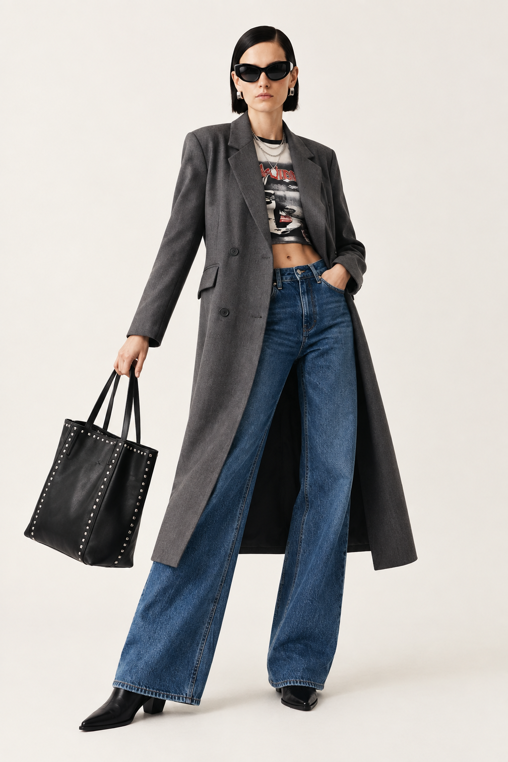 A model stands against a light beige studio background wearing a long charcoal gray double-breasted blazer over a cropped graphic tee, blue wide-leg jeans, black pointed-toe heels, and black sunglasses, while holding a black studded leather tote bag.