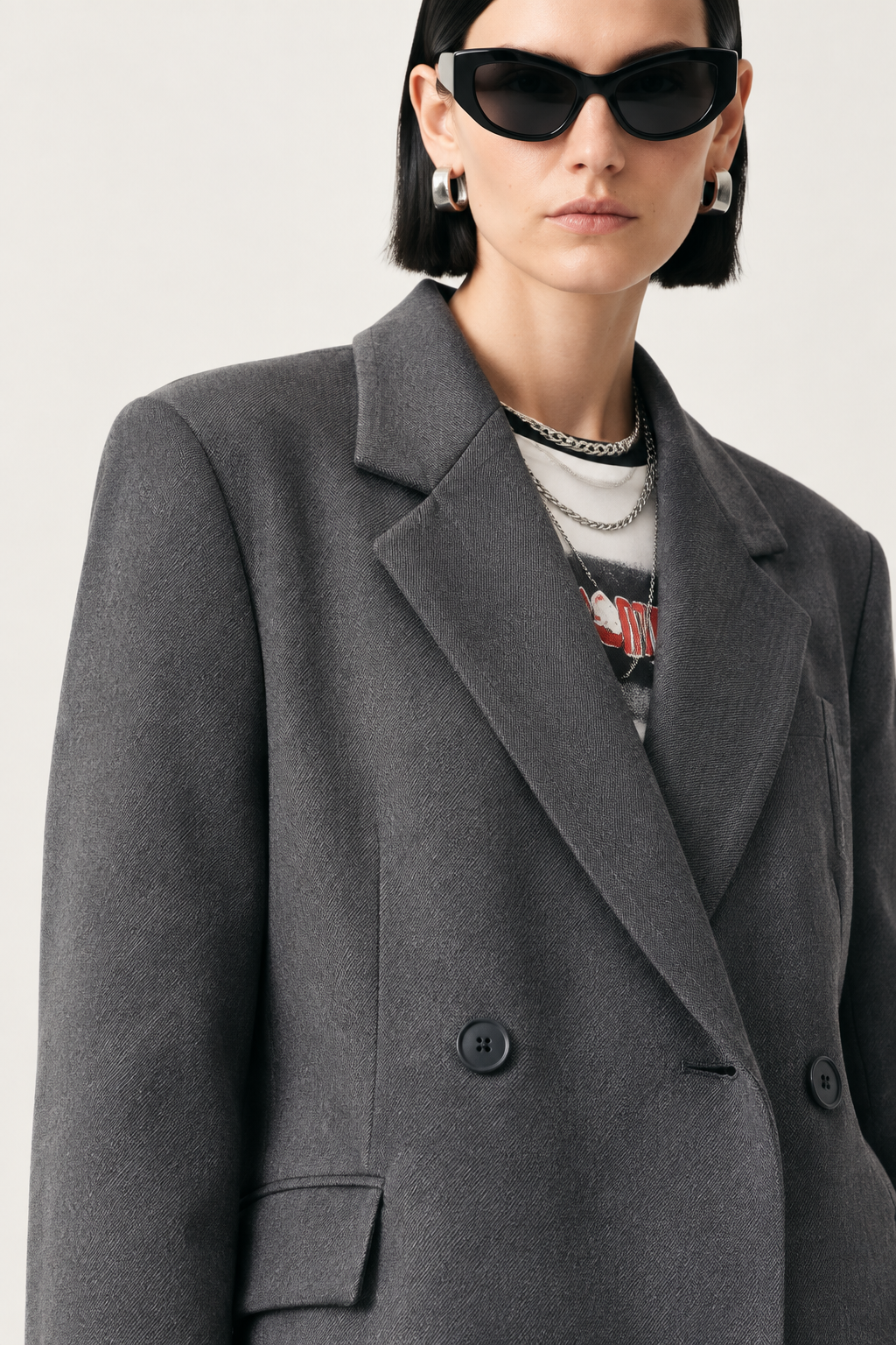 A close-up of a model wearing a dark gray double-breasted blazer over a graphic t-shirt, accessorized with black cat-eye sunglasses, layered silver necklaces, and silver hoop earrings against a light gray background.