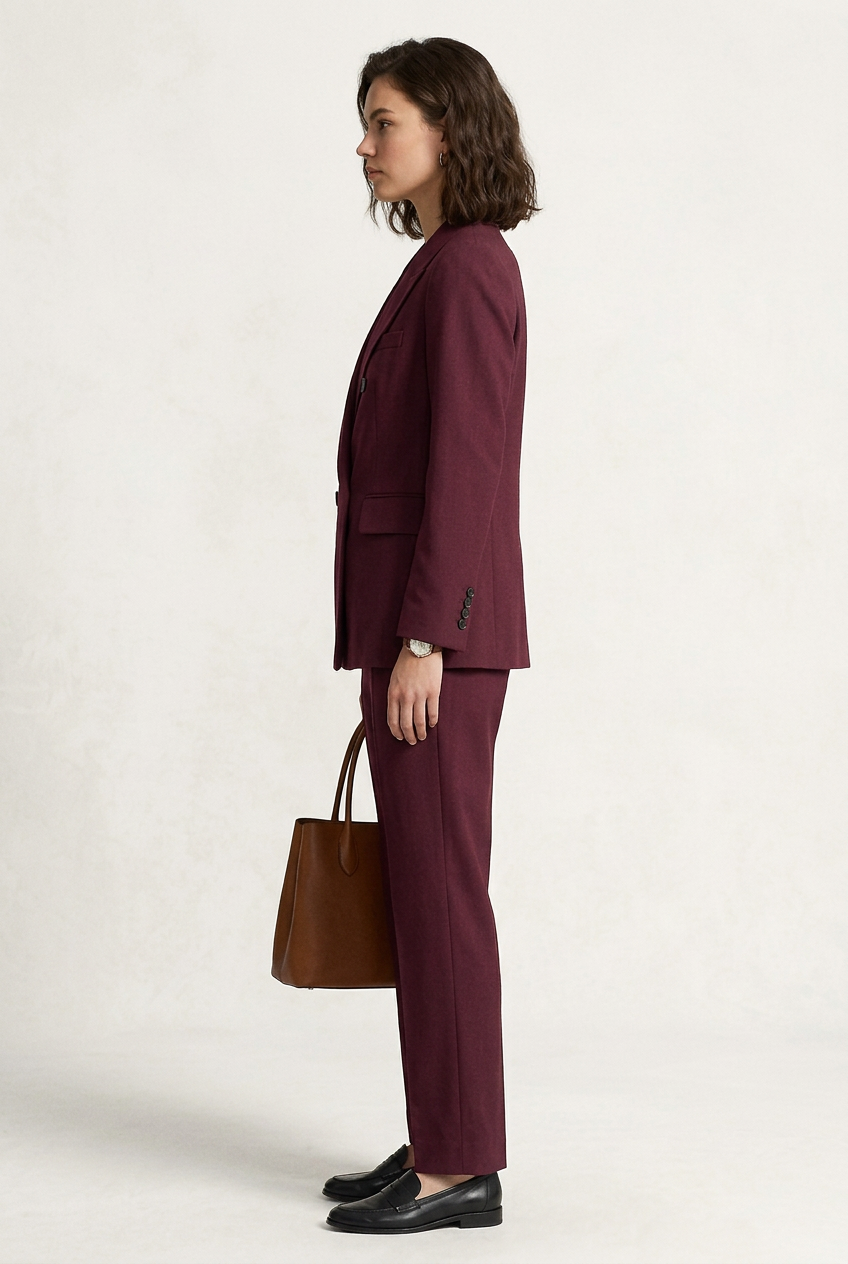A woman models a matching burgundy double-breasted blazer and trousers suit, holding a brown leather handbag and wearing black loafers, against a plain off-white studio background.