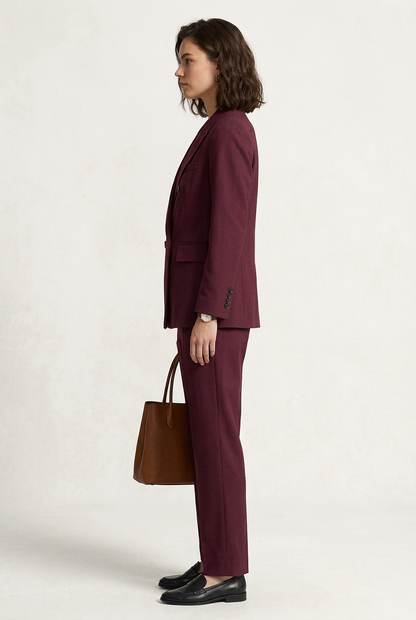 A woman models a matching burgundy double-breasted blazer and trousers suit, holding a brown leather handbag and wearing black loafers, against a plain off-white studio background.
