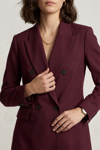 A woman models a matching burgundy double-breasted blazer and trousers suit, holding a brown leather handbag and wearing black loafers, against a plain off-white studio background.