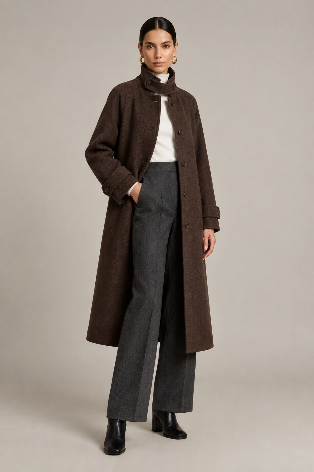 A model stands against a plain light gray studio background wearing a long dark brown coat over a white top and gray wide-leg trousers, with black ankle boots.