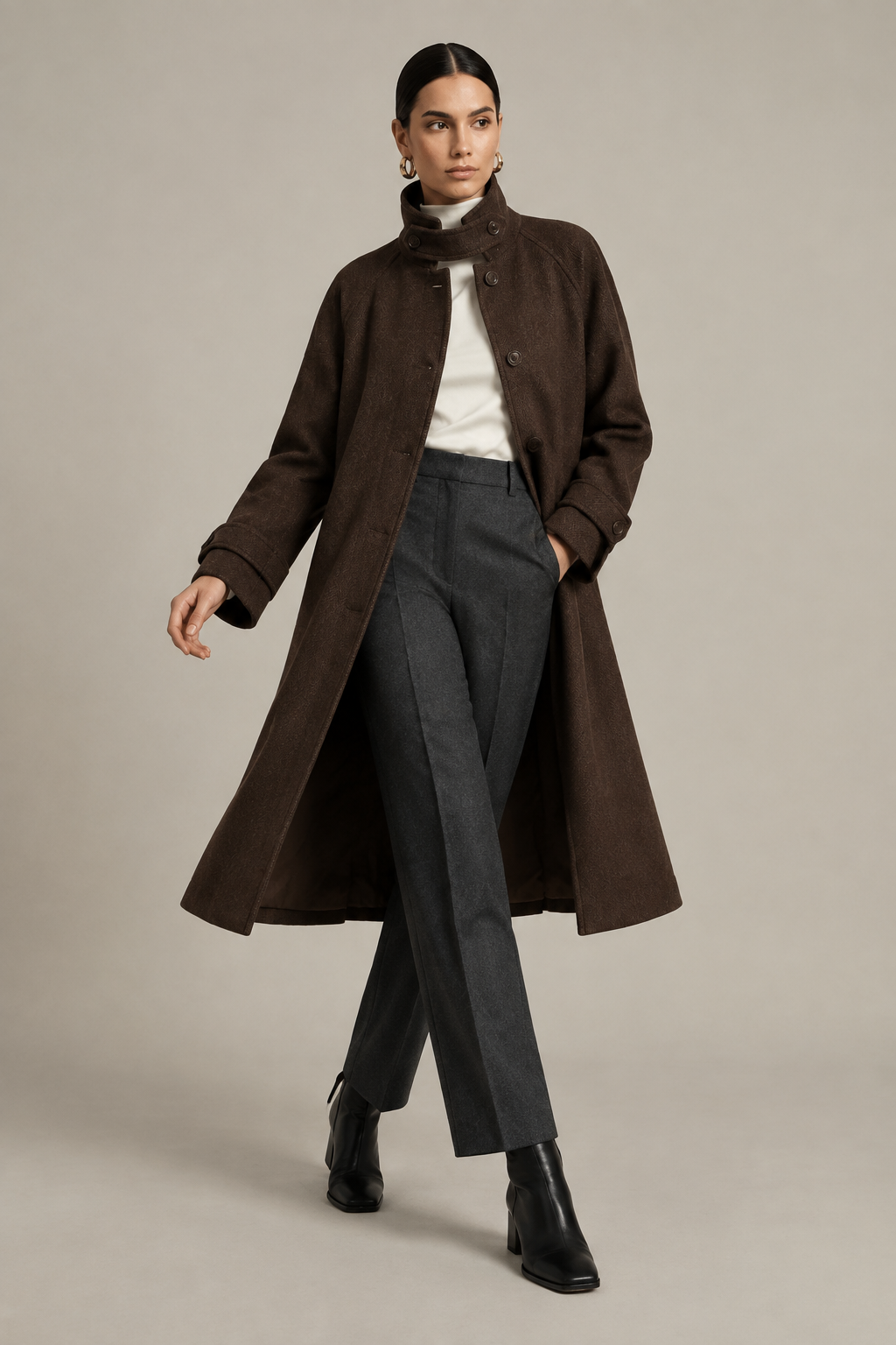 A model is shown wearing a long brown wool-blend coat with a high mock neck, styled with a white turtleneck, dark gray trousers, and black ankle boots, set against a plain light gray studio background.