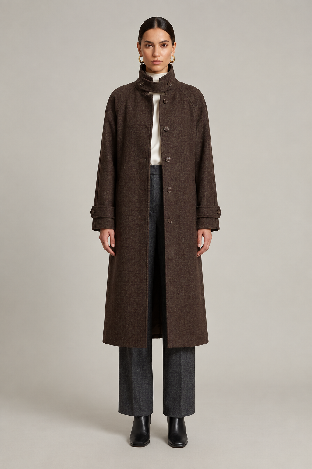 A model stands against a neutral light gray studio background wearing a long dark brown coat over a white top, dark gray wide-leg trousers, and black ankle boots.
