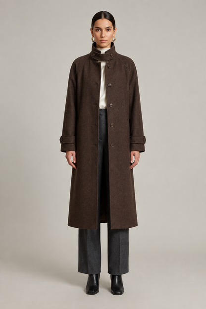 A model stands against a neutral light gray studio background wearing a long dark brown coat over a white top, dark gray wide-leg trousers, and black ankle boots.