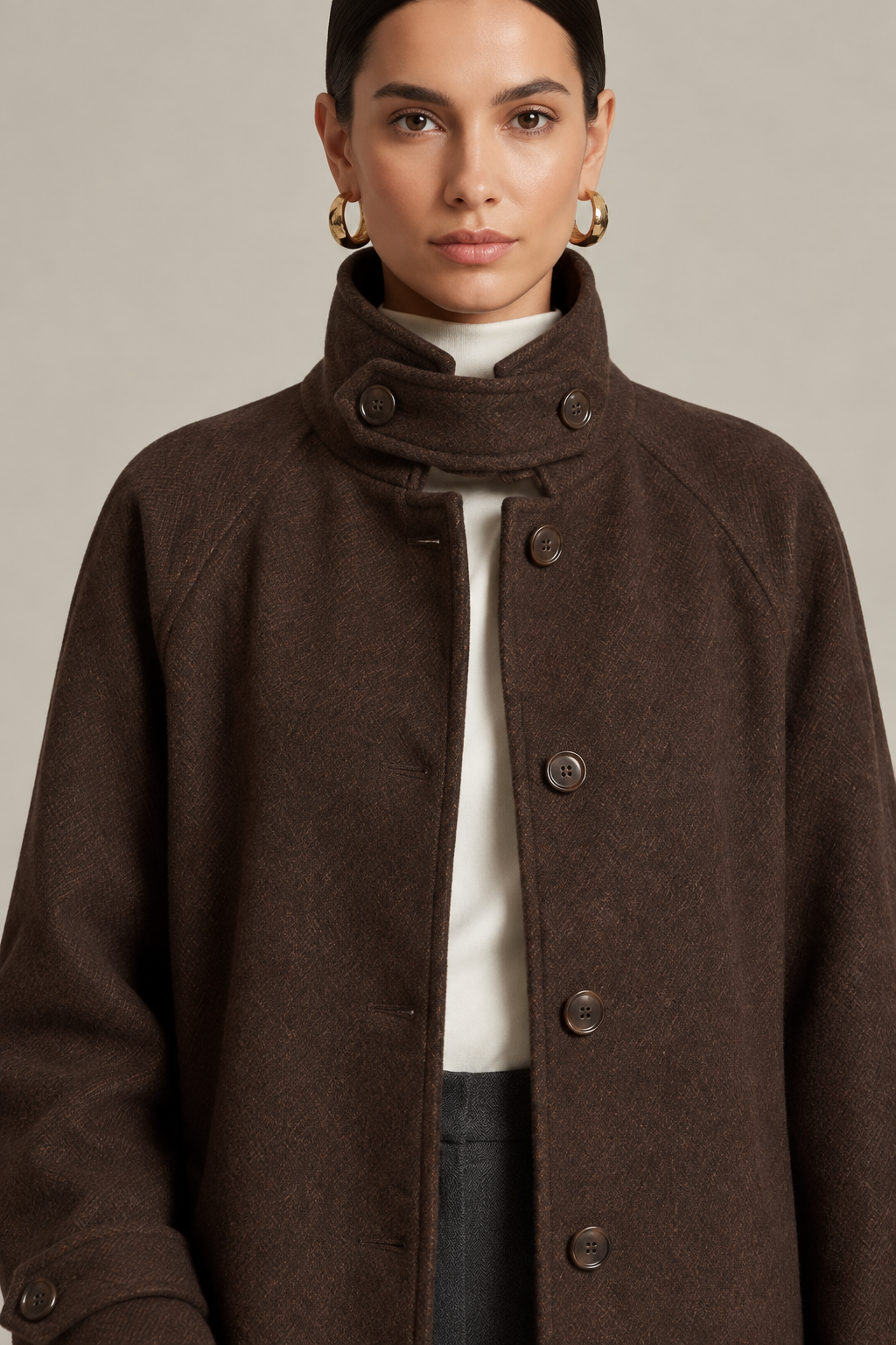 A close-up of a model wearing a dark brown textured wool coat with a high mock neck and large dark buttons, layered over a white turtleneck and dark trousers.