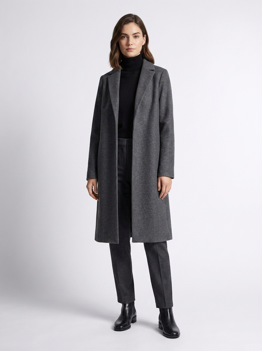 A woman modeling a long charcoal gray wool-blend coat over a black turtleneck, dark gray trousers, and black ankle boots, standing against a light gray studio background.