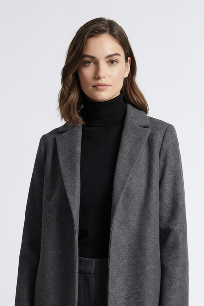 A woman modeling a long charcoal gray wool-blend coat over a black turtleneck, dark gray trousers, and black ankle boots, standing against a light gray studio background.